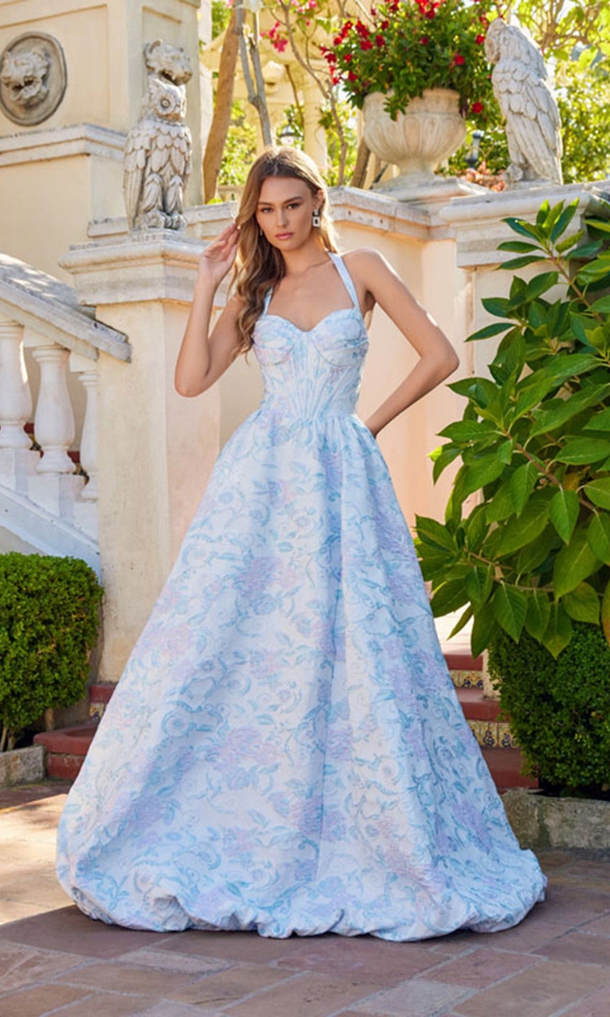 A woman stands outside among stone statues and greenery, wearing the Juliet JT2742A Long Prom Dress in Aqua Mist, featuring a fitted corset bodice, thin straps, and floral-textured fabric.